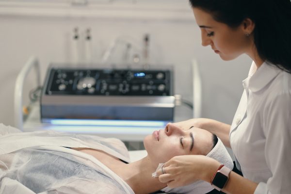 Woman lying during cosmetic procedure while skillful female cosmetologist making facial treatment